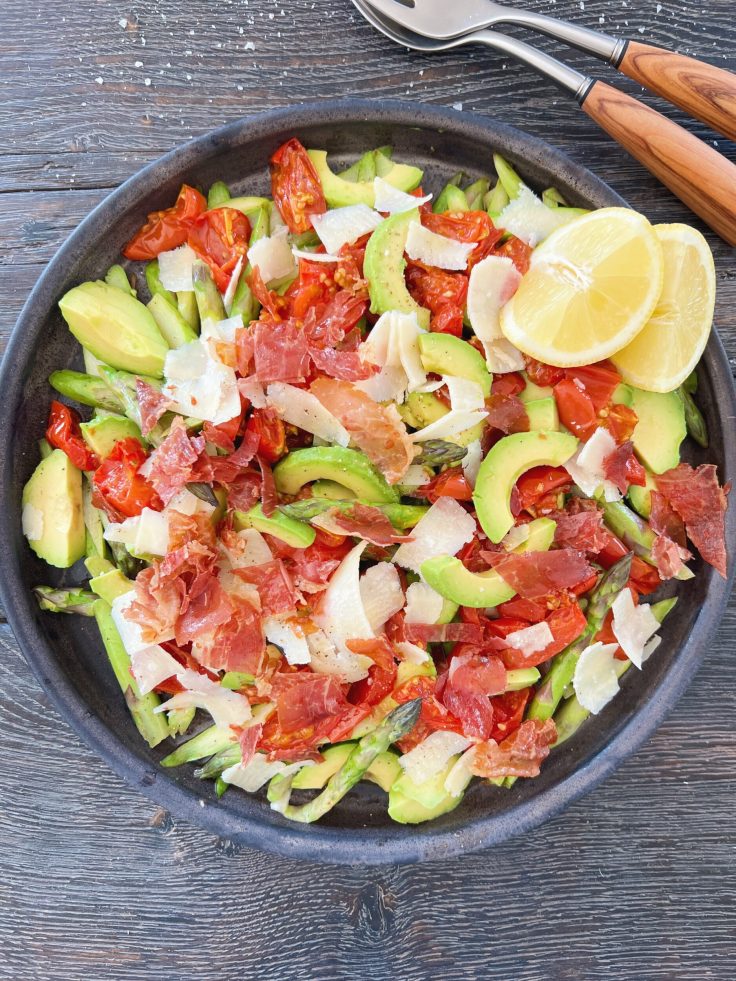 salat med råstegte asparges blandet med ovnbagte tomater avocado og parmesan serveret i keramikfad der er sort og er fra brandet badass. Ved siden af salaten ligger der et salatbestik med skaft i oliventræ