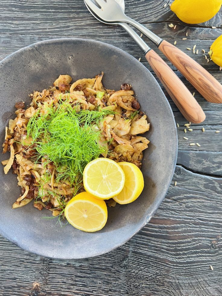 En mørk og rustik keramikskål står på et sort rustikt træbord med en gylden grøntsagsret. Stegt fennikel med grøn toppien og der er placeret halve citroner i den ene side. Ved siden af skålen ligger der et salatbestik med håndtag i oliventræ